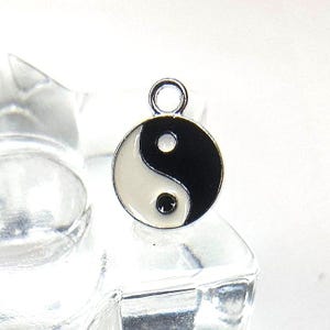 以下が含まれることがあります： エナメル加工が施された黒と白の陰陽シンボルが描かれた円形の金属製チャーム。チャームの上部には取り付け用の小さな銀色のループがあり、対照的な色の2つの小さな点を持つバランスの取れたクラシックなデザインが特徴です。
