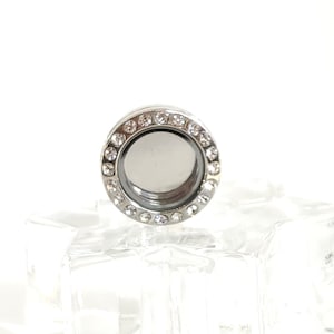 May include: A silver-toned locket with a circular design, encrusted with small, clear crystals around the outer edge. The locket has a clear, reflective center. The locket is displayed on a clear, faceted glass block against a white background.
