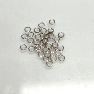 May include: A collection of small, circular silver-toned metal jump rings. Each individual ring features a thin wire construction with a visible split or opening, designed for connecting jewelry components or other small craft items together.