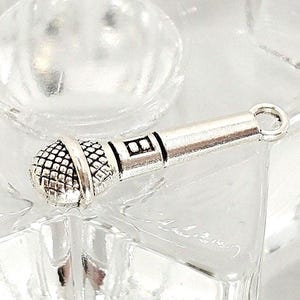 May include: A silver-colored microphone charm with a detailed mesh head and a cylindrical body. The charm has a small loop at the top for attaching to a necklace or bracelet. The microphone is resting on a clear, textured surface.