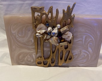 May include: A rectangular, pearlescent clutch with a swirling pattern. The gold-toned clasp features an abstract design adorned with crystals, pearls, and iridescent shell fragments. The clutch is a neutral color, suitable for various outfits.