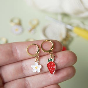 Könnte beinhalten: Zwei goldfarbene Maschenmarkierer mit Anhängern. Ein Anhänger ist eine weiße Blume mit gelber Mitte. Der andere Anhänger ist eine rote Erdbeere mit grünen Blättern.