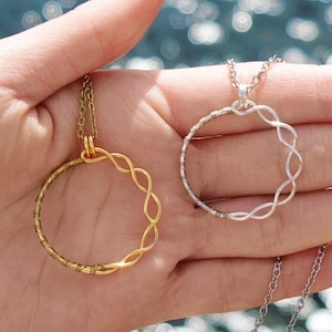 May include: Two silver and gold necklaces with a circle pendant design. The necklaces are on a hand with a blurred background of water.