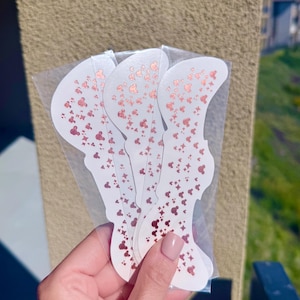 Puede incluir: Un conjunto de pegatinas cosméticas blancas, troqueladas con un patrón oro rosa de estrellas y siluetas de Mickey Mouse. Las pegatinas están en una funda de plástico transparente. El fondo es una pared neutra y soleada.