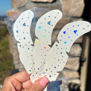 Könnte beinhalten: Drei weiße Aufkleber in Federform mit holografischem Glanz und kleinen Akzenten in Form von Mickey Mouse-Köpfen und Sternen. Die Aufkleber befinden sich in einer transparenten Kunststoffverpackung.