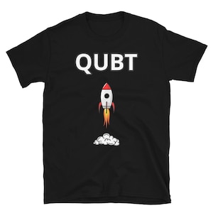 Può includere: T-shirt nera con la scritta "QUBT" in bianco sopra un razzo dei cartoni animati. Il razzo è bianco con la punta rossa e fiammate arancioni. Fumo bianco sotto il razzo suggerisce il decollo.