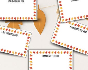 May include: White cards with the text "I AM THANKFUL FOR" and "I AM GRATEFUL FOR" are displayed. Each card is bordered with a decorative leaf pattern in red, yellow, and orange. The cards are hanging with paper leaf cutouts.