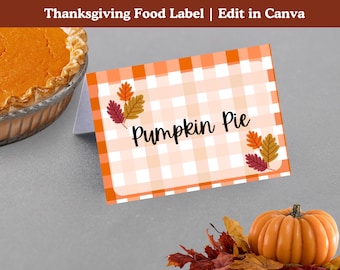 May include: Thanksgiving food label with the text "Pumpkin Pie" on a white and orange checkered background. The label is decorated with fall leaves. A pumpkin pie and a small orange pumpkin are also visible.
