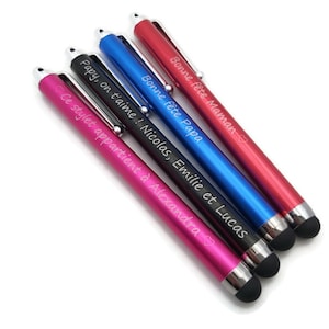 Puede incluir: Cinco bolígrafos estilizados personalizados con cuerpos de diferentes colores y mensajes grabados. Los bolígrafos son negros, rojos, azules, rosas y plateados. Los mensajes son "Bonne fête Maman", "Bonne fête Papa", "Papy, on t'aime! Nicolas, Emilie et Lucas", "Ce stylet appartient à Alexandra" y un símbolo de corazón.