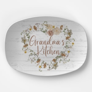 May include: Oval serving platter with a white wood-look background. A floral wreath in neutral tones encircles the words "Grandma's Kitchen" in a warm brown script. The flowers include white, yellow, and brown accents.