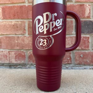May include: A maroon stainless steel tumbler with a handle and a clear plastic lid. The front features the white Dr Pepper logo, the text Est. 1885, and a circular emblem containing the number 23 with the text Authentic Blend of 23 Flavors.
