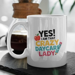 May include: White ceramic mug with the text "YES! I AM THAT CRAZY DAYCARE LADY" in colourful letters. The design includes a red apple and a pencil. The mug has a large handle and is sitting on a black surface.