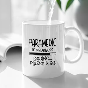 Può includere: Tazza in ceramica bianca con la scritta "PARAMEDIC in progress LOADING... PLEASE WAIT" in nero. L'acqua viene versata nella tazza. Una pianta verde è sullo sfondo.