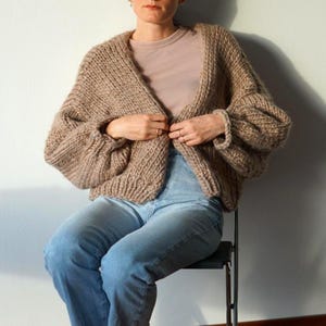 Può includere: Un cardigan a maglia grossa di colore beige neutro. Il maglione aperto ha un ampio colletto a coste e maniche voluminose. La modella indossa il cardigan con un top rosa chiaro e jeans blu. Il cardigan è realizzato con un filato spesso e morbido.