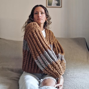 Può includere: Un cardigan a maglia grossa nei toni del marrone e del grigio. Il maglione oversize presenta una trama a coste e maniche ampie. L'indumento è indossato sopra un top bianco e jeans chiari. Il cardigan è un capo comodo ed elegante.