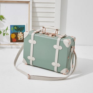 May include: A light green vintage-style suitcase with white trim and gold accents. The suitcase has a top handle and a long strap. The suitcase is closed and has a lock on the front.