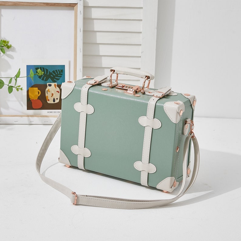 May include: A light green vintage-style suitcase with white trim and gold accents. The suitcase has a top handle and a long strap. The suitcase is closed and has a lock on the front.