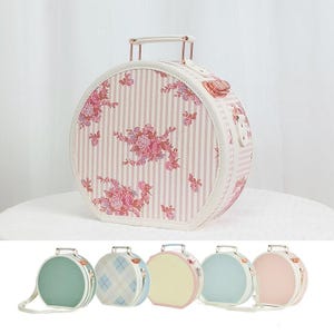 May include: A round, cream-colored suitcase with a pink floral design and vertical stripes. The suitcase has a handle and a clasp closure. Additional suitcases in various pastel colors are displayed below.