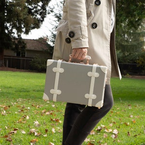 May include: A gray and white vintage-style suitcase with gold accents and white straps. The suitcase is being held by a person wearing a beige trench coat.