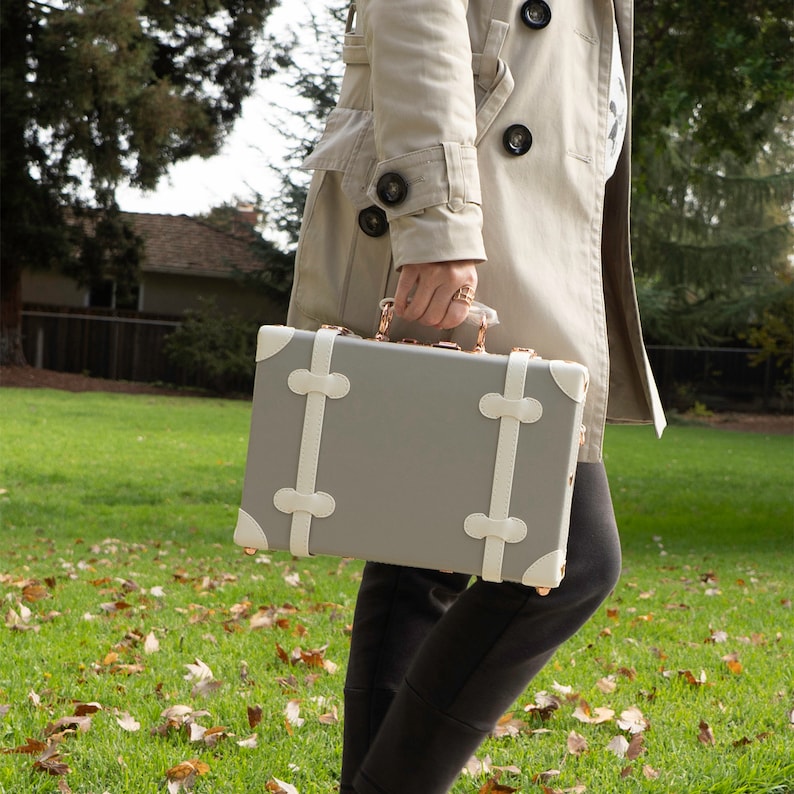 May include: A gray and white vintage-style suitcase with gold accents and white straps. The suitcase is being held by a person wearing a beige trench coat.