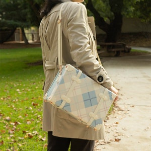 May include: A beige trench coat with a plaid patterned suitcase attached to the shoulder strap. The suitcase has a light blue and pink plaid pattern with gold accents.