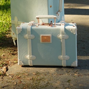 May include: A light blue vintage-style suitcase with white leather accents and a rose pattern. The suitcase has a brass lock and handle.