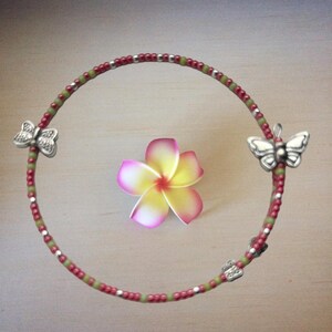 May include: A beaded bracelet with red, green, and silver beads, featuring three silver butterfly charms. A pink and yellow flower rests in the center of the bracelet, creating a decorative arrangement.