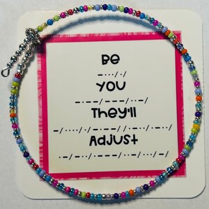 May include: A colorful beaded anklet with a silver clasp. The beads are in various colors, including blue, pink, green, and orange. A card behind the anklet reads "Be You, They'll Adjust" in Morse code.