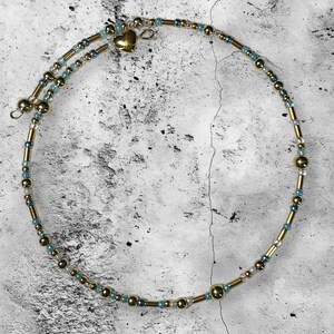 May include: A delicate necklace featuring a spiral design. The necklace is adorned with gold-colored beads, small blue beads, and a small gold heart pendant. The necklace is designed to be worn around the neck.
