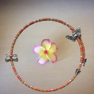 May include: A beaded bracelet with orange, green, and silver beads, adorned with three silver butterfly charms. A pink, yellow, and white flower rests in the center, creating a nature-inspired theme.