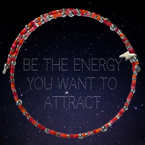 May include: A wrap bracelet with red, orange, and pink beads, interspersed with silver-colored beads. A small, white star charm is attached. The bracelet is displayed against a starry night background with the text "BE THE ENERGY YOU WANT TO ATTRACT."