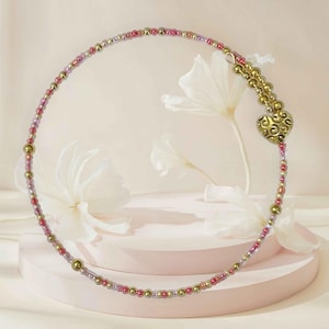 May include: A delicate necklace featuring a heart-shaped pendant and a beaded design. The necklace is adorned with small, colorful beads in shades of pink, gold, and purple. The heart pendant has an ornate, swirling pattern.