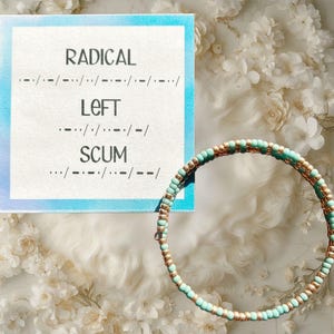 May include: A beaded bracelet with turquoise and gold-colored beads. The bracelet is coiled and sits next to a card with the words "RADICAL LEFT SCUM" in Morse code. The card has a blue border.