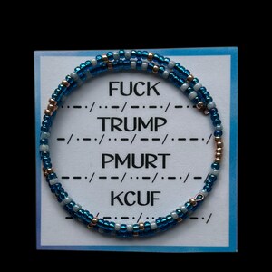 May include: A beaded bracelet with blue, white, and gold beads is displayed on a white card with a blue border. The card has the words "FUCK TRUMP PMURT KCUF" and Morse code symbols.