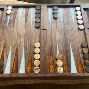 May include: A handcrafted wooden backgammon board with a rich, dark wood finish. The board features inlaid resin accents in shades of white and amber, and includes wooden game pieces and dice. A small owl figurine is carved into the corner.