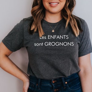 May include: A woman wearing a dark gray t-shirt with the text "Les ENFANTS sont GROGNONS" in white. She is wearing blue jeans with a button fly.
