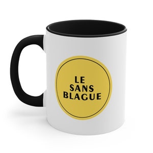 May include: White ceramic mug with a black rim and handle. The mug features a yellow circle with the text "LE SANS BLAGUE" in black.