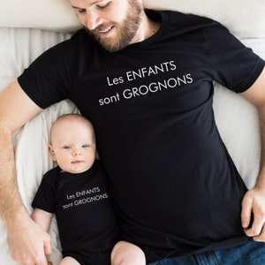 May include: Black t-shirt and onesie set with the text "Les ENFANTS sont GROGNONS" in white. The t-shirt is worn by an adult, and the onesie is worn by a baby. The image is taken from above.
