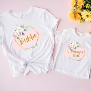May include: Two white t-shirts with floral designs and gold accents. The larger shirt says "Bubbe" and the smaller shirt says "Bubbe's girl".