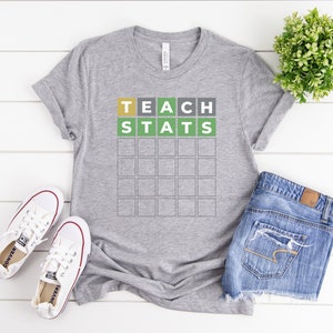May include: A gray t-shirt with the words "TEACH STATS" in a Wordle-style design. The shirt is paired with white sneakers and denim shorts. A small green plant is visible in the background.