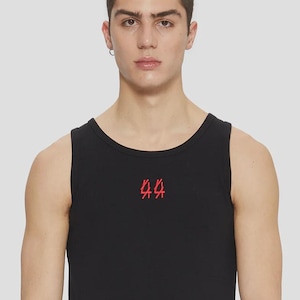 May include: Black tank top with a red graphic on the chest. The sleeveless shirt is made of a soft material and has a rounded neckline. The model is wearing black pants.
