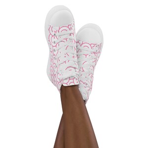Peut inclure: Baskets montantes blanches avec un motif rose r&eacute;p&eacute;titif. Le motif est compos&eacute; de demi-cercles roses. Les baskets ont des lacets blancs et une semelle en caoutchouc blanche. Les chaussures sont pr&eacute;sent&eacute;es avec les pieds crois&eacute;s.