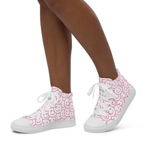 Peut inclure: Baskets montantes blanches avec un motif r&eacute;p&eacute;t&eacute; de visages souriants roses. Les chaussures ont des lacets blancs et une semelle blanche. Les baskets en toile pr&eacute;sentent un design amusant et ludique.