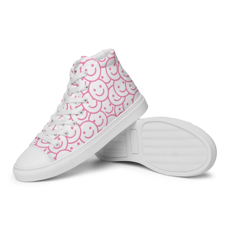 Peut inclure: Baskets montantes blanches avec un motif r&eacute;p&eacute;t&eacute; de visages souriants roses. Les chaussures ont des lacets blancs et une semelle blanche. Le design est ludique et amusant.