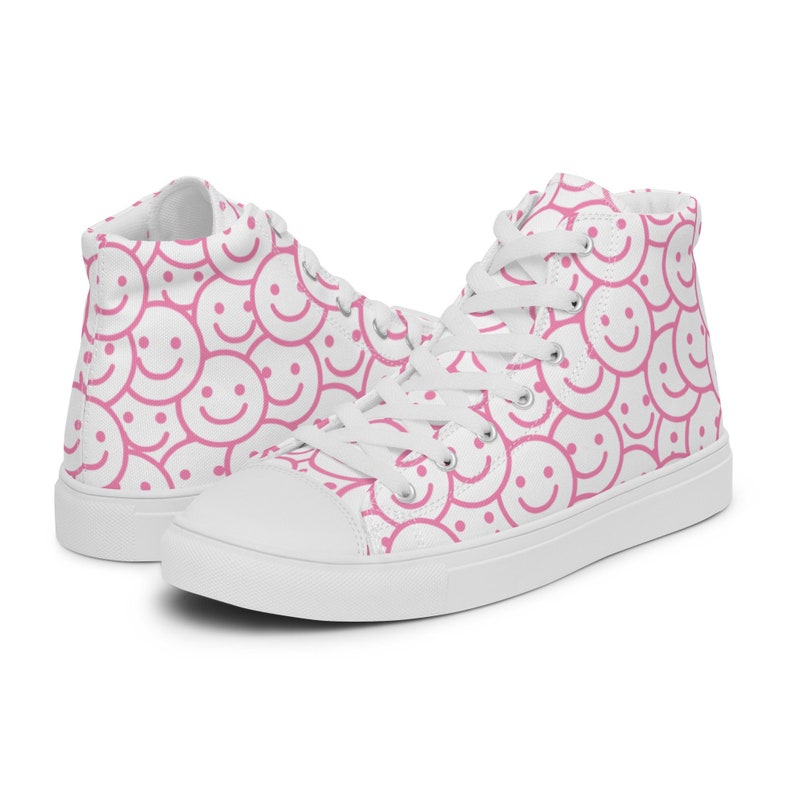 Peut inclure: Baskets montantes blanches avec un motif r&eacute;p&eacute;t&eacute; de visages souriants roses. Les chaussures ont des lacets blancs et une semelle en caoutchouc blanche. Le mat&eacute;riau en toile est recouvert du motif joyeux.