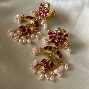 Peut inclure: Une paire de boucles d'oreilles dorées avec un clou en forme de lotus et une pampille en forme de croissant, incrustées de pierres rouge foncé et de cristaux clairs. Les boucles sont ornées de perles d'eau douce blanches suspendues.