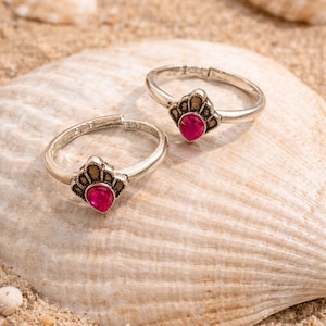 Peut inclure: Deux bagues argentées dotées d'un anneau ouvert et d'un sertissage décoratif en forme d'éventail. Chaque bague est ornée d'une pierre précieuse rose vif en forme de goutte. Le métal présente une finition légèrement oxydée et détaillée.