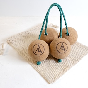 Può includere: Quattro palline da massaggio in sughero con manico e dettagli in corda verde acqua, ciascuna con un logo circolare nero. Le palline poggiano su un sacchetto di rete beige. Progettate per l'automassaggio e il sollievo muscolare.