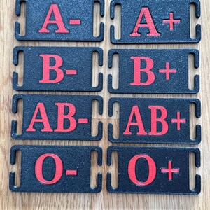 May include: Eight black rectangular tags with red lettering indicating blood types: A-, A+, B-, B+, AB-, AB+, O-, and O+. Each tag has a slot on each end.