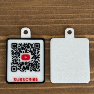 May include: Two white rectangular tags with a hole at the top. One tag features a black and white QR code with a red YouTube logo and the word "SUBSCRIBE" in red. The other tag is plain white.